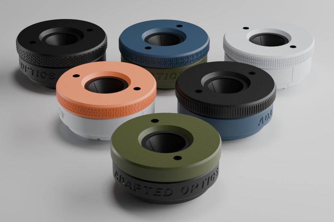 Six cylindrical hardware components or rings in assorted colors, including black, blue, white, orange, olive green, and dark gray, arranged on a light gray studio background.